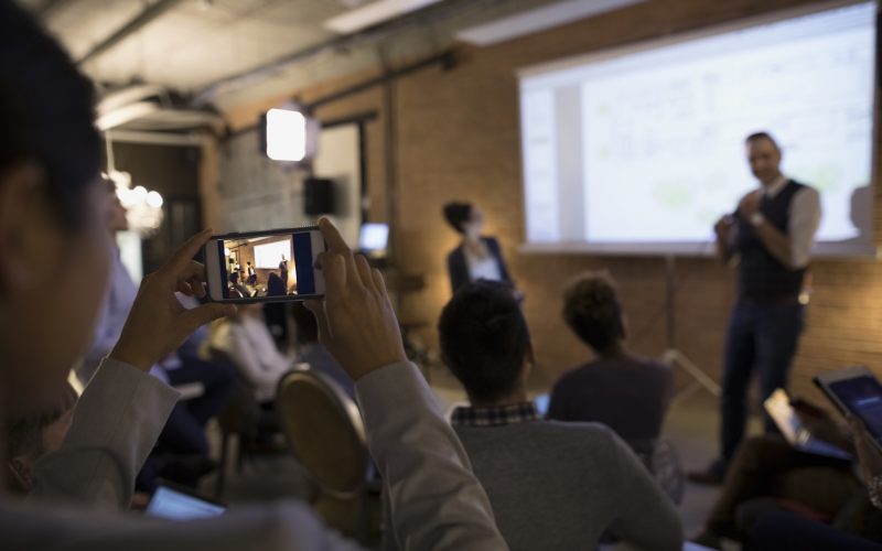 Businesswoman with camera phone videoing conference speaker in conference room